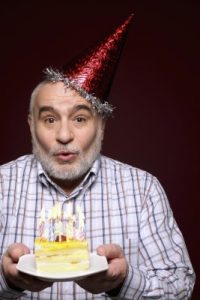 an older man with a birthday hat on blowing out candles on his birthday cake.