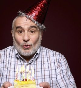 an older man with a birthday hat on blowing out candles on his birthday cake.