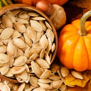 Close up image of toasted, salted pumpkin seeds spilling from a wooden bowl, accented with nuts and a small pumpkin
