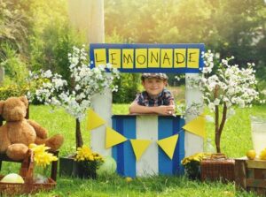 a little boy has an outdoor homemade lemonade stand with a sign and he looks happy for a small business or money concept