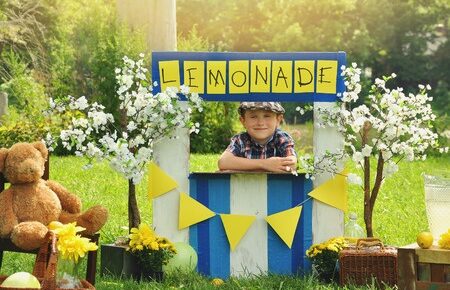 a little boy has an outdoor homemade lemonade stand with a sign and he looks happy for a small business or money concept